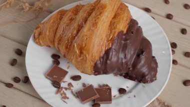 Kahve çekirdekleri ve kurumuş çiçeklerle beyaz bir tabakta çikolatalı kruvasanın sinematik yakın çekimi. Pürüzsüz kamera hareketi desen, fırın havası, kahvaltı veya kafe kavramını vurgular