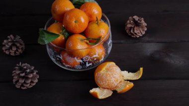 Koyu ahşap bir masada içi taze mandalinalarla dolu cam bir Noel kasesi. Soyulmuş mandalina ve kozalak sıcak bir kış tatili ortamı yaratır. Şenlikli mevsimlik gıda geçmişi