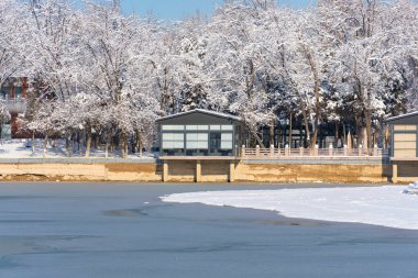 Kış kenti parkında buzun pürüzsüz olduğu donmuş bir göl. Karla kaplı ağaçlar, açık mavi gökyüzü ve soğuk güneşli bir günde sakin bir atmosfer. Mevsimlik kış manzarası
