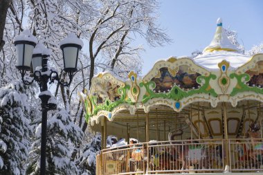 Karlı bir parkta hayvanlarla ve süslü panellerle süslenmiş parlak bir atlıkarınca duruyor. Canlı renkler ve dingin kış manzarası.