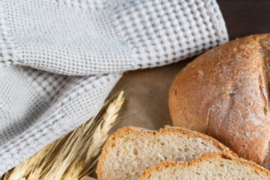 Taze pişmiş rustik ekmek somunu ahşap yüzeyde kısmen dilimlenmiş, buğday sapları ve bez ile eşleştirilmiş, sağlıklı bir çiftlikten sofraya hissi için.
