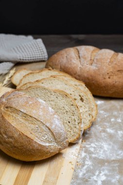 Taze pişmiş rustik ekmek somunu ahşap yüzeyde kısmen dilimlenmiş, buğday sapları ve bez ile eşleştirilmiş, sağlıklı bir çiftlikten sofraya hissi için.
