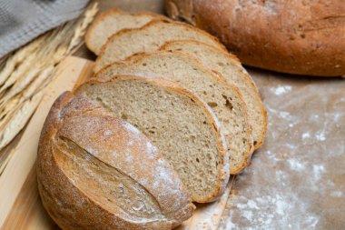 Taze pişmiş rustik ekmek somunu ahşap yüzeyde kısmen dilimlenmiş, buğday sapları ve bez ile eşleştirilmiş, sağlıklı bir çiftlikten sofraya hissi için.