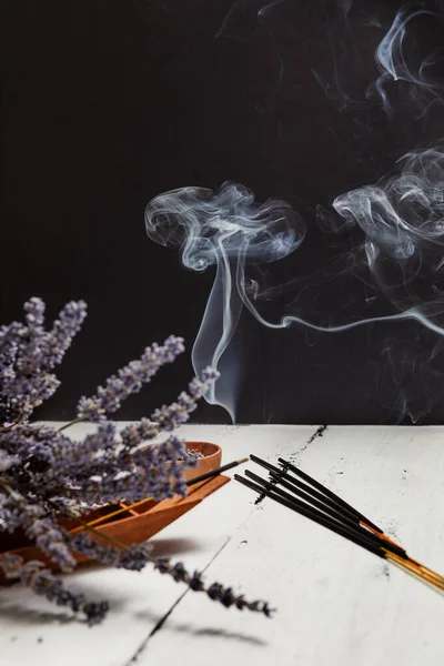 Asian incense sticks and a bouquet of dried lavender on a white wooden table against a dark wall, a wave of incense. Peaceful atmosphere.