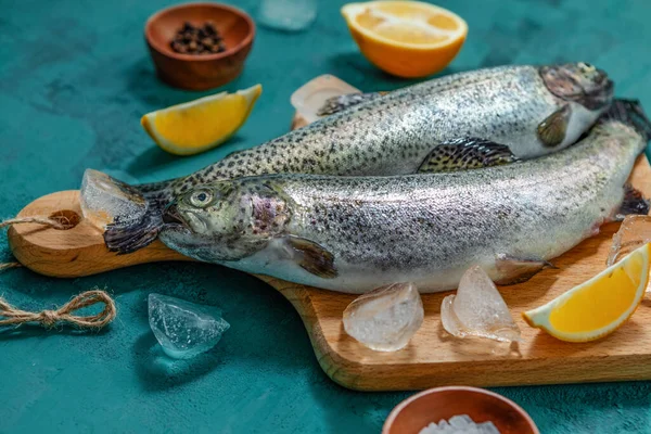 Two fish, raw trout with ice, spices and lemon on a cutting Board on a turquoise background. close-up