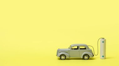 Gri oyuncak bir araba ve sarı arka planda bir pil. Elektrikli araçlar, ekoloji ve iklim koruma kavramı