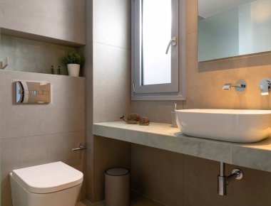 Modern tarz banyo içi pencereli gri taş döşeme, kap lavabo, ayna arka lambası, duvara monte edilmiş tuvalet.