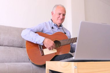 Mature man with headphones learning to play guitar online with laptop alone at home. Concept learning within covid19 pandemics. clipart