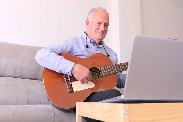 自宅でノートパソコンだけでオンラインでギターを演奏することを学ぶヘッドフォンを持つ成熟した男 Covid19流行病における概念学習 ストック画像