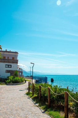 Açık ve güneşli bir günde, sakin mavi deniz boyunca uzanan, bulutların arasından bakan uzak dağlarla ve her iki taraftaki yeşilliklerle dolu pitoresk bir yürüyüş yolu. Sapri, Campania, İtalya.