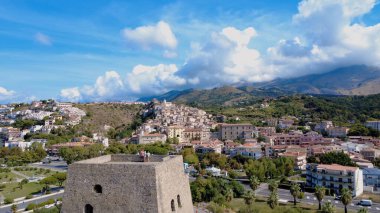 Aşağıda renkli binaları olan bir sahil kasabası uzanırken, ön planda tarihi bir taş kule duruyor. Dağlar parlak mavi bir gökyüzünün altında yükselir. Scalea, Calabria, İtalya.