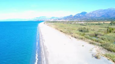 Güneşli bir günde, arka planda berrak su ve dağlarla uzun bir sahili keşfetmek.