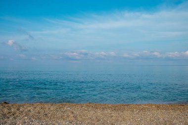 Yumuşak bulutlar ufukta süzülürken berrak su ufuk boyunca uzanıyor. Kıyı küçük çakıl taşlarıyla kaplı. Gün ışığında doğal bir plaj sahnesi yaratıyor..