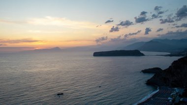 Güneş okyanusun üzerinde batıyor, suyun üzerine sıcak bir parıltı saçıyor. Arka planda dağlar yükselir ve bulutlar gökyüzünde sürüklenir. Kıyı şeridi deniz boyunca uzanıyor..