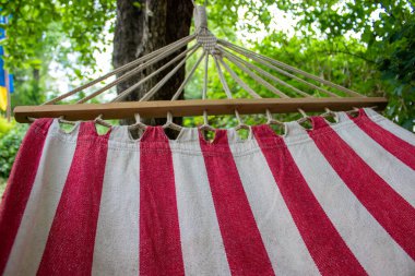 Yeşil yapraklar ve benekli güneş ışığıyla çevrili kırmızı ve beyaz çizgili bir hamak bir ağaca asılır. Sahnede güneşli bir günde rahatlama ve açık hava eğlencesi gösteriliyor..