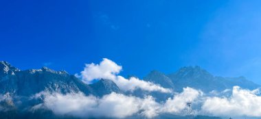 Bulutlar dağları kaplarken açık mavi bir gökyüzü sahneyi dolduruyor. Kar dağın tepelerinde dinleniyor, yakındaki bir köy için zemin hazırlıyor..