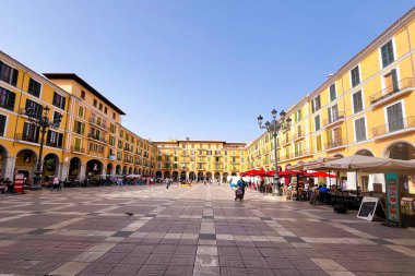 Palma, İspanya, 30 Ekim 2024: İnsanlar sarı binalarla çevrili geniş bir meydanda yürüyorlar. Masalar ve sandalyeler dışarıda yemek için hazır. Gökyüzü açık, parlak bir atmosfer yaratıyor..