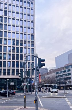 Frankfurt am Main, Almanya, Kasım 02, 2024: Otobüsler trafik ışığında beklerken insanlar bir şehir kavşağında karşıdan karşıya geçiyor. Gökyüzü bulutlu ve bölgeyi çevreleyen birkaç yüksek bina var..