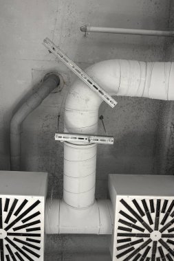 Bir binanın tavanında borular ve havalandırmalar görülüyor. Manzara havalandırma sisteminin düzenini gösteren yapıya bakıyor. Bölge iyi aydınlatılmış..