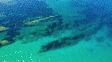 Parlak mavi su, kıyı bölgesindeki su altındaki kayaları ve deniz yaşamını ortaya çıkarıyor. Güneş ışığı, okyanus tabanının net görüş açısını gösteren yansımalar yaratır..