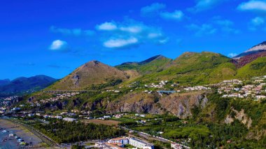 Hava manzarası dağları ve yeşil ağaçların arasına yuva yapmış evleri gösteriyor. Gökyüzü çok az bulutla aydınlık ve aşağısı çok canlı..