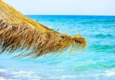 Sahildeki altın kamış plaj şemsiyesinin yakın çekimi, arka planda mavi dalgalı su olan manzara, yaz yaşam tarzı