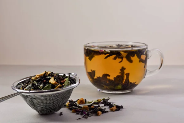 Glass, transparent cup with hot brewed tea, metal strainer filter with a dry mix of herbs and fruits on a light wooden background. Concept for a tea ceremony. Close up.