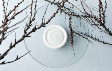 Açık bir krema kavanozuyla kompozisyon, cam üzerinde kuru dallar, mavi arka plan. Üst Manzara. Soyut. 