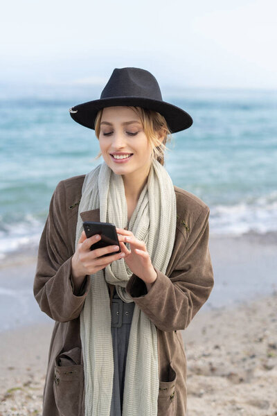 Stylish young woman scrolling a feed on the mobile phone with a smile on the beach 