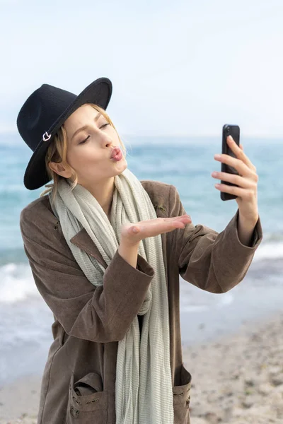 Stylish happy girl sending a kiss through a video call using her mobile hone on a beach