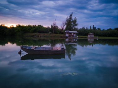 Bulutlu bir yaz akşamı Sava nehrinin manzarası, sakin sulardaki bulutların yansıması, diğer kıyıdaki balıkçı teknesi ve balıkçı kulübeleri.