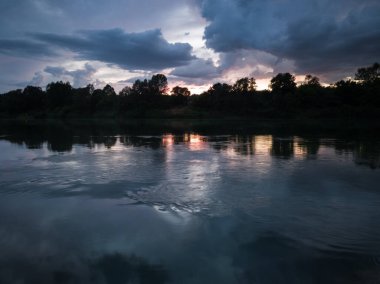 Nehir manzarası, bulutlar ve günbatımında orman kıyıları. Mavi saatte bulutlu alacakaranlıkta sakin nehir manzarası