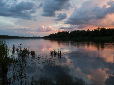 Gün batımında nehir, bulutlar, su çimleri ve dağ manzarası. Bulutlu alacakaranlıkta son güneş bulutlarda parlarken huzurlu nehir manzarası