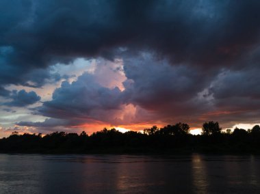 Nehir manzarası, karanlık bulutlar ve gün batımında orman kıyıları. Bulutlu havalarda dramatik nehir manzarası. Yağmur veya yaz yağmuru ve bulutlarda muhteşem kırmızı-turuncu bir parıltı.