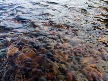 Altında renkli çakıl taşları olan sığ, dalgalı bir akarsu yüzeyi derede akan su, dalgalanan suyla samimi bir manzara.