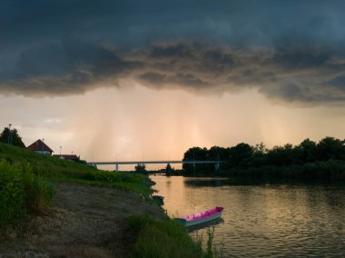 Şiddetli yağış veya yaz yağmuru, şiddetli hava ve yağmurun ardında parlayan güneş fırtınası ve kümülonimbus bulutu. Çimenli nehir kıyısı ve köprü boyunca demirli teknelerle Sava nehri manzarası.