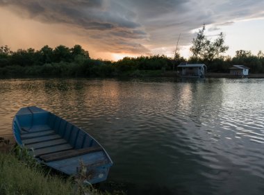 Şiddetli yağış veya yaz yağmuru, şiddetli hava ve yağmurun ardında parlayan güneş fırtınası ve kümülonimbus bulutu. Çimenli nehir kıyısı ve kulübe boyunca demirli tekneyle Sava nehri manzarası.