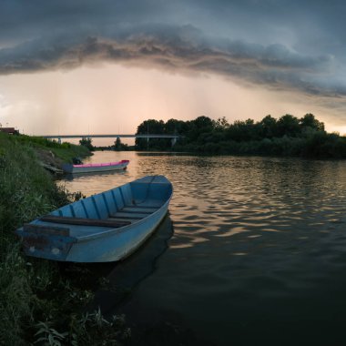 Şiddetli yağış veya yaz yağmuru, şiddetli hava ve yağmurun ardında parlayan güneş fırtınası ve kümülonimbus bulutu. Çimenli nehir kıyısı ve köprü boyunca demirli teknelerle Sava nehri manzarası.