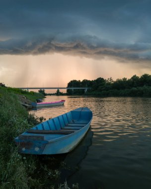 Şiddetli yağış veya yaz yağmuru, şiddetli hava ve yağmurun ardında parlayan güneş fırtınası ve kümülonimbus bulutu. Çimenli nehir kıyısı ve köprü boyunca demirli teknelerle Sava nehri manzarası.