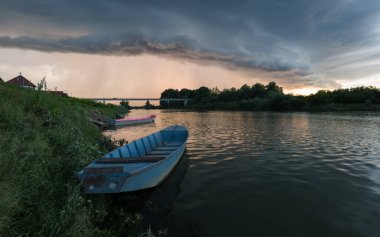 Şiddetli yağış veya yaz yağmuru, şiddetli hava ve yağmurun ardında parlayan güneş fırtınası ve kümülonimbus bulutu. Çimenli nehir kıyısı ve köprü boyunca demirli teknelerle Sava nehri manzarası.