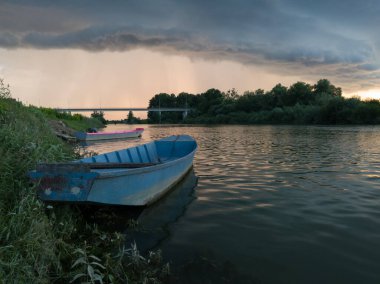 Şiddetli yağış veya yaz yağmuru, şiddetli hava ve yağmurun ardında parlayan güneş fırtınası ve kümülonimbus bulutu. Çimenli nehir kıyısı ve köprü boyunca demirli teknelerle Sava nehri manzarası.