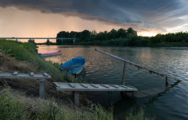 Şiddetli yağış veya yaz yağmuru, şiddetli hava ve yağmurun ardında parlayan güneş fırtınası ve kümülonimbus bulutu. Tahta rıhtım ve köprünün yanında demirli teknelerle Sava nehri manzarası