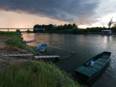 Şiddetli yağış veya yaz yağmuru, şiddetli hava ve yağmurun ardında parlayan güneş fırtınası ve kümülonimbus bulutu. Tahta rıhtım ve köprünün yanında demirli teknelerle Sava nehri manzarası