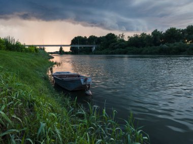 Şiddetli yağış veya yaz yağmuru, şiddetli hava ve yağmurun ardında parlayan güneş fırtınası ve kümülonimbus bulutu. Çimenli kıyısı, demirli teknesi ve köprüsü olan Sava nehri manzarası.