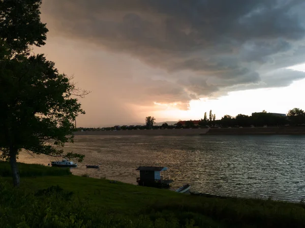 Storm cumulonimbus cloud with heavy rain or summer shower, severe weather and sun glow behind rain. Landscape with Sava river and moored boats in Bosanski Brod, Bosnia and Herzegovina