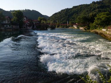 Banja Luka yakınlarındaki Novoselija 'daki Vrbas nehrinde şelale. Güneşli bir yaz akşamında nehir, köy ve orman dağları manzarası.