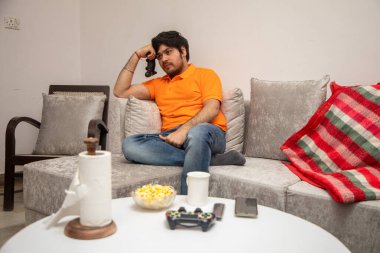 Asyalı Hintli adam oyun oynuyor, canlı yayın yapıyor ve TV seyrediyor. Oturma odasında, gri bir kanepe ve beyaz bir masa üzerinde..