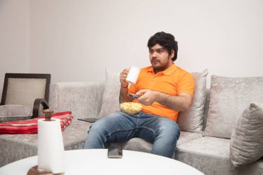 Asyalı Hintli adam oyun oynuyor, canlı yayın yapıyor ve TV seyrediyor. Oturma odasında, gri bir kanepe ve beyaz bir masa üzerinde..