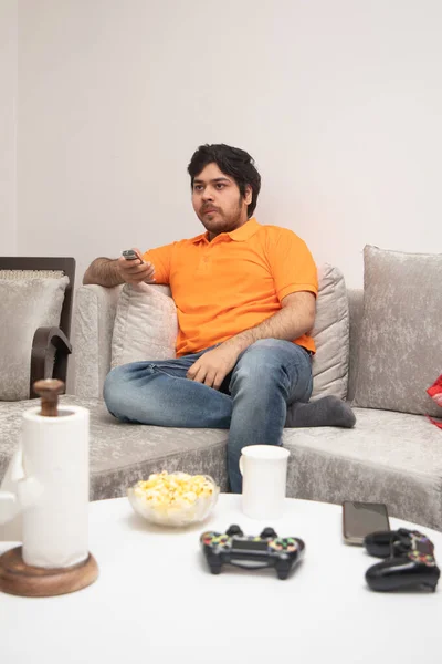 Asian Indian man gaming, streaming and watching TV alone in living room on a grey sofa and white table.