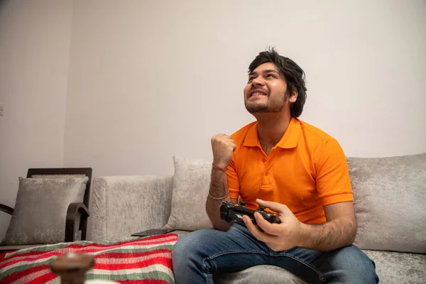 Asyalı Hintli adam oyun oynuyor, canlı yayın yapıyor ve TV seyrediyor. Oturma odasında, gri bir kanepe ve beyaz bir masa üzerinde..